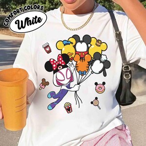 Puede incluir: Camiseta blanca con un gr&aacute;fico colorido que presenta un personaje de dibujos animados sosteniendo globos con forma de personajes de Disney. La camiseta tiene las palabras "COMFORT COLORS" y "White" en un dise&ntilde;o circular. Iconos de comida de dibujos animados adicionales.