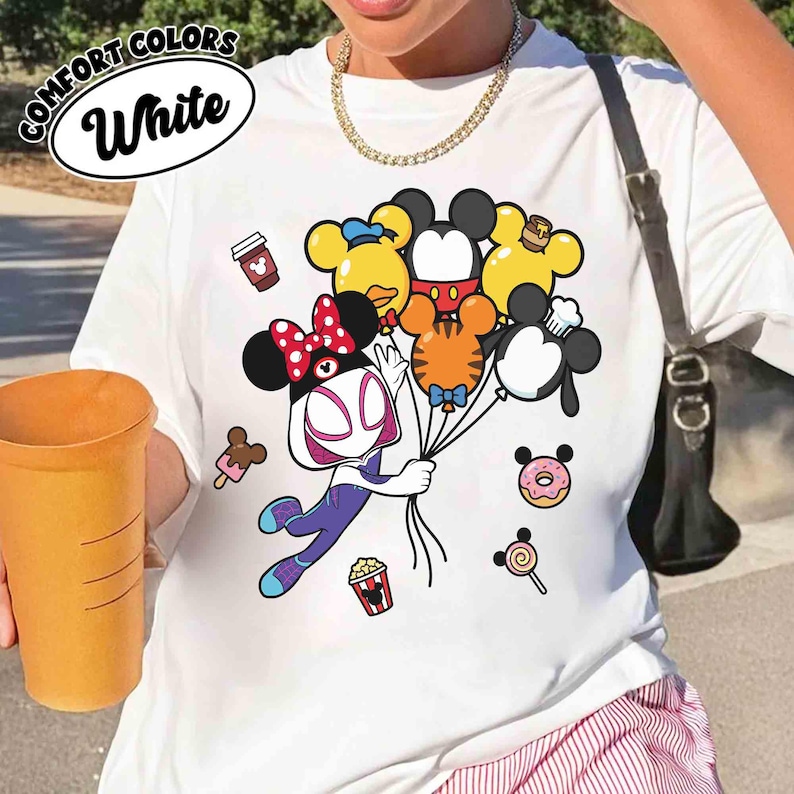 Puede incluir: Camiseta blanca con un gr&aacute;fico colorido que presenta un personaje de dibujos animados sosteniendo globos con forma de personajes de Disney. La camiseta tiene las palabras "COMFORT COLORS" y "White" en un dise&ntilde;o circular. Iconos de comida de dibujos animados adicionales.