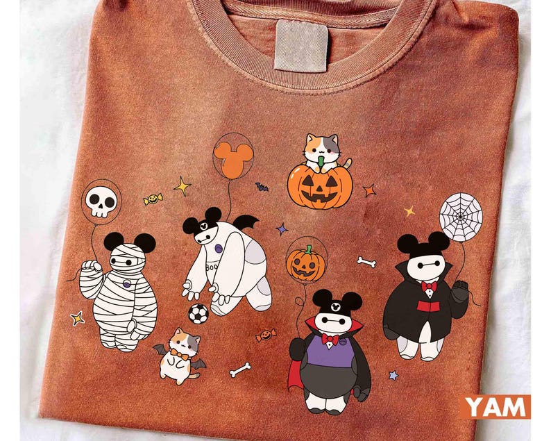 May include: Rust-colored t-shirt featuring cartoon characters dressed in Halloween costumes. Characters include a mummy, vampire, and Baymax-inspired figures, along with a cat in a pumpkin. The design includes Halloween-themed elements like candy, stars, and a spiderweb.