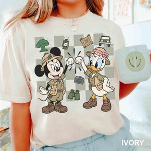 May include: Ivory-colored t-shirt featuring a cartoon illustration of Mickey Mouse and Daisy Duck in safari outfits, giving a fist bump. The design includes safari-themed icons like a jeep, map, and binoculars. A white mug with a smiley face is also visible.
