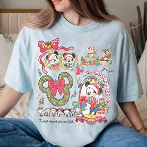 May include: Light blue t-shirt featuring a festive Disney Christmas design. The graphic includes Mickey Mouse, Minnie Mouse, Chip and Dale, and other characters, along with the text "Very Merry Christmas 2025" and "The most magical place on Earth."