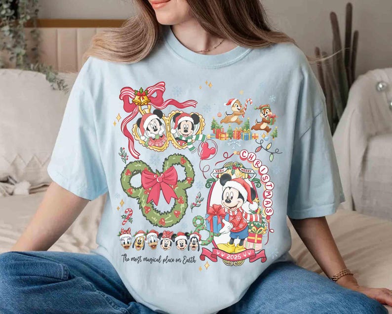 May include: Light blue t-shirt featuring a festive Disney Christmas design. The graphic includes Mickey Mouse, Minnie Mouse, Chip and Dale, and other characters, along with the text "Very Merry Christmas 2025" and "The most magical place on Earth."