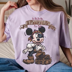 Puede incluir: Camiseta morada con una imagen de dibujos animados de Mickey Mouse vestido de pirata. El texto de la camiseta dice "Yo Ho, Yo Ho, A Pirates Life For Me!"