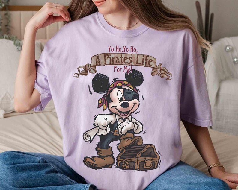 Puede incluir: Camiseta morada con una imagen de dibujos animados de Mickey Mouse vestido de pirata. El texto de la camiseta dice "Yo Ho, Yo Ho, A Pirates Life For Me!"