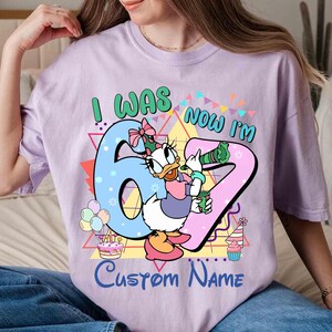 May include: A light purple t-shirt with the text "I WAS 6 NOW I'M 7" and "Custom Name". The shirt features a cartoon image of Daisy Duck holding a drink, with birthday decorations.
