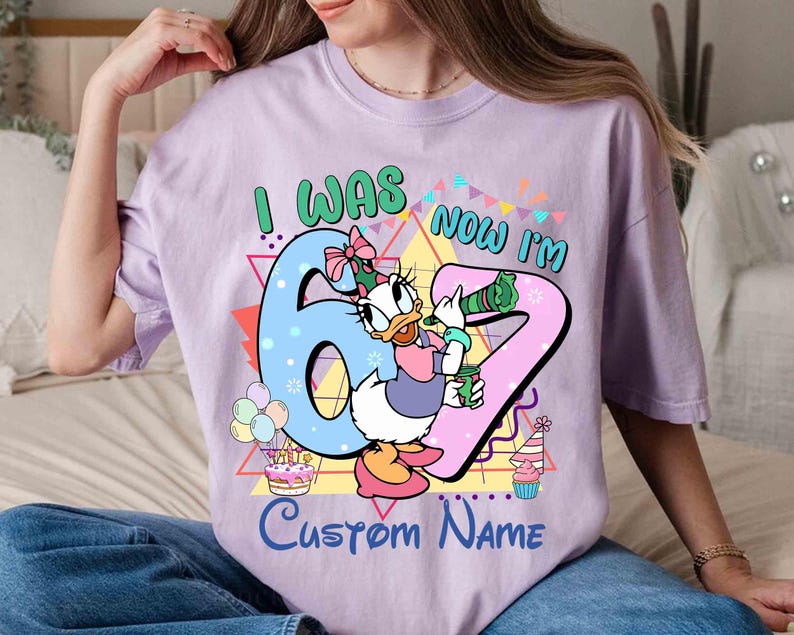 May include: A light purple t-shirt with the text "I WAS 6 NOW I'M 7" and "Custom Name". The shirt features a cartoon image of Daisy Duck holding a drink, with birthday decorations.