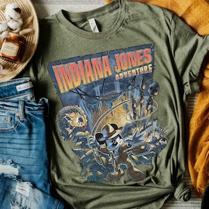 May include: A green t-shirt with a graphic print of Mickey Mouse dressed as Indiana Jones. The text "Indiana Jones Adventure" is printed on the shirt.