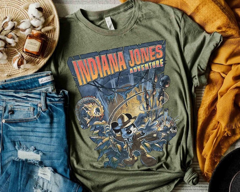 May include: A green t-shirt with a graphic print of Mickey Mouse dressed as Indiana Jones. The text "Indiana Jones Adventure" is printed on the shirt.