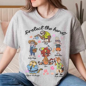 Peut inclure: T-shirt gris avec un imprim&eacute; graphique de personnages du film Zootopie. Le texte "Prot&eacute;gez le troupeau !" est imprim&eacute; sur le t-shirt. Les personnages incluent Judy Hopps, Nick Wilde, Flash, Yax et Bellwether.