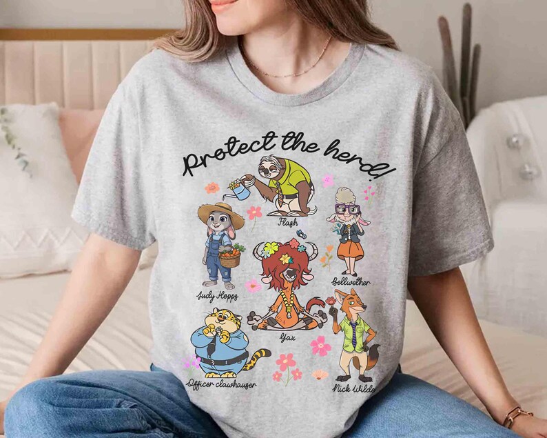 Peut inclure: T-shirt gris avec un imprim&eacute; graphique de personnages du film Zootopie. Le texte "Prot&eacute;gez le troupeau !" est imprim&eacute; sur le t-shirt. Les personnages incluent Judy Hopps, Nick Wilde, Flash, Yax et Bellwether.