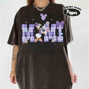 Puede incluir: Camiseta negra con un gr&aacute;fico de Daisy Duck sosteniendo una flor y una cuchara. La palabra "MIMI" est&aacute; en letras de cuadros morados. La camiseta tambi&eacute;n tiene un lazo morado, corazones y el texto "Comfort Colors Pepper".