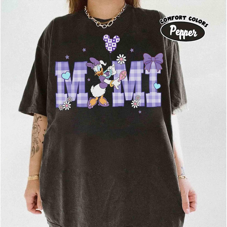 Puede incluir: Camiseta negra con un gr&aacute;fico de Daisy Duck sosteniendo una flor y una cuchara. La palabra "MIMI" est&aacute; en letras de cuadros morados. La camiseta tambi&eacute;n tiene un lazo morado, corazones y el texto "Comfort Colors Pepper".