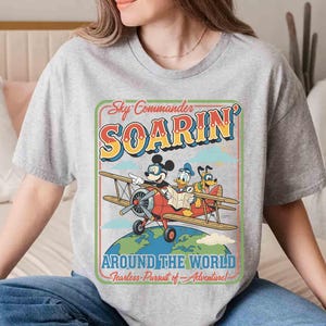 May include: A gray t-shirt featuring a vintage-style graphic of Mickey Mouse, Donald Duck, and Goofy in a biplane. The design includes the text "Sky Commander Soarin' Around The World" in a retro font, with the tagline "Fearless Pursuit of Adventure!"