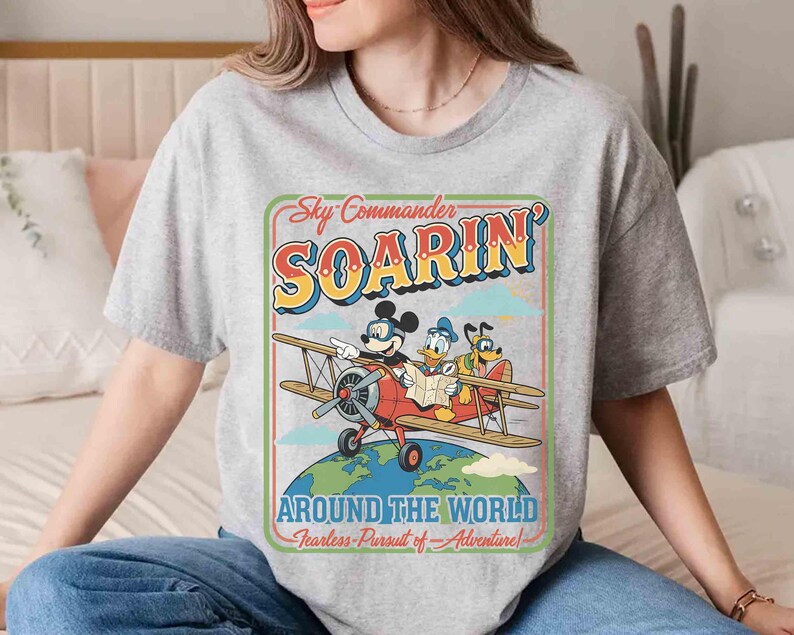 May include: A gray t-shirt featuring a vintage-style graphic of Mickey Mouse, Donald Duck, and Goofy in a biplane. The design includes the text "Sky Commander Soarin' Around The World" in a retro font, with the tagline "Fearless Pursuit of Adventure!"