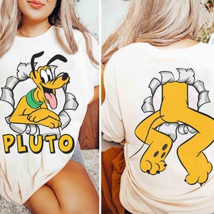 May include: Cream-coloured t-shirt with a cartoon Pluto graphic. The front shows Pluto emerging from a torn circle, with the name "PLUTO" below. The back displays Pluto's legs and paws. A fun, playful design.