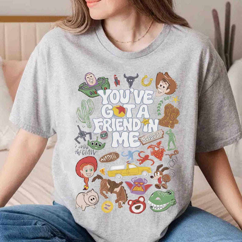 May include: A light gray t-shirt featuring a colorful graphic design with characters and text from the movie "Toy Story". The text reads "You've Got a Friend in Me". The design includes cartoon images of Woody, Buzz Lightyear, and other characters.