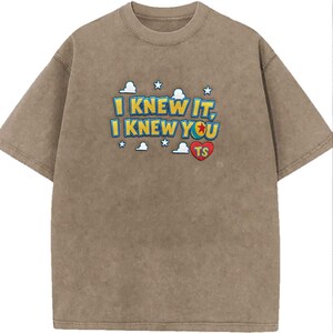 May include: A brown short-sleeved t-shirt with a vintage wash effect. The chest features embroidered text that reads I KNEW IT, I KNEW YOU with a red heart containing the letters TS, surrounded by small white cloud and star icons in blue and yellow.