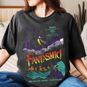 K&ouml;nnte beinhalten: Dunkelgraues T-Shirt mit farbenfrohem Grafikdesign. Das Design zeigt einen lila Drachen, das Wort "FANTASMIC!" in Rot und Gelb und den Text "GOOD CLASHES WITH EVIL IN A NIGHTTIME SPECTACULAR".