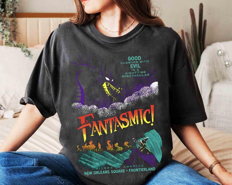 K&ouml;nnte beinhalten: Dunkelgraues T-Shirt mit farbenfrohem Grafikdesign. Das Design zeigt einen lila Drachen, das Wort "FANTASMIC!" in Rot und Gelb und den Text "GOOD CLASHES WITH EVIL IN A NIGHTTIME SPECTACULAR".