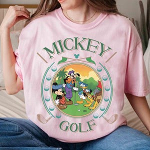 May include: Pink t-shirt featuring a circular graphic of Mickey Mouse, Goofy, Donald Duck, and Pluto playing golf. The words "MICKEY GOLF" are arched above and below the image in green. Golf clubs and golf balls are also featured.