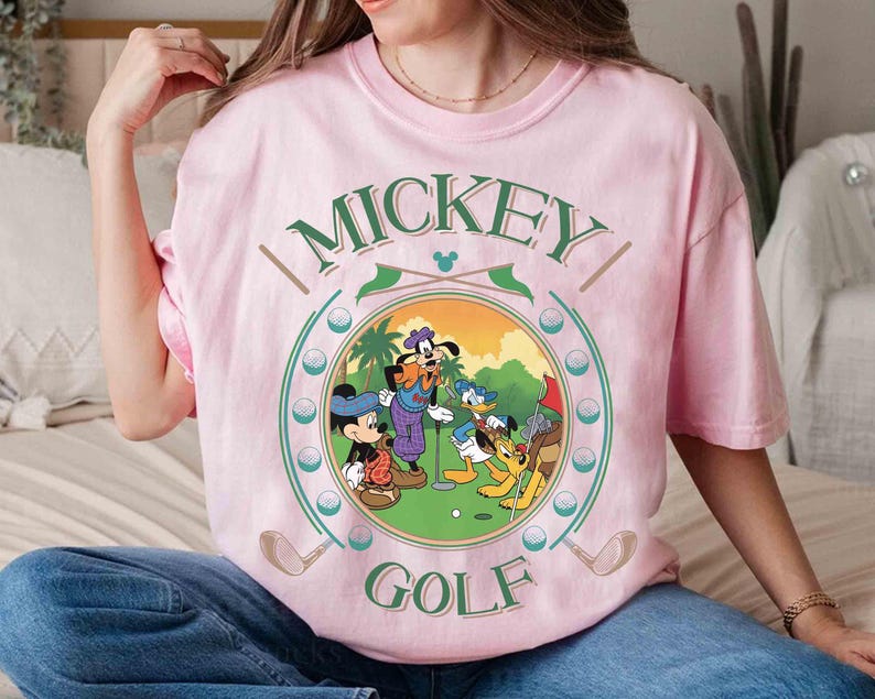 May include: Pink t-shirt featuring a circular graphic of Mickey Mouse, Goofy, Donald Duck, and Pluto playing golf. The words "MICKEY GOLF" are arched above and below the image in green. Golf clubs and golf balls are also featured.