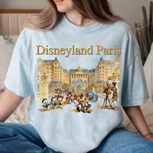 May include: Light blue t-shirt featuring the text "Disneyland Paris" in gold lettering above a colorful illustration of Disney characters in front of a grand building. The shirt has short sleeves and a relaxed fit.