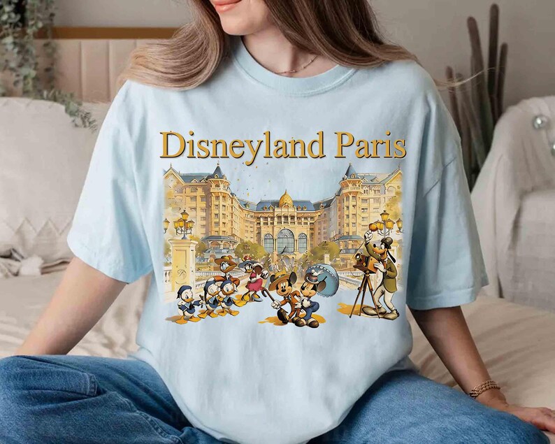May include: Light blue t-shirt featuring the text "Disneyland Paris" in gold lettering above a colorful illustration of Disney characters in front of a grand building. The shirt has short sleeves and a relaxed fit.