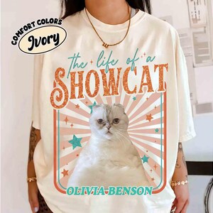 Może przedstawiać: Kremowa koszulka z napisem "the life of a SHOWCAT" w kolorze pomarańczowym i brokatowymi literami. Ilustracja kota jest wyśrodkowana, z retro wzorem z gwiazdami i paskami. Poniżej znajduje się imię "OLIVIA-BENSON".