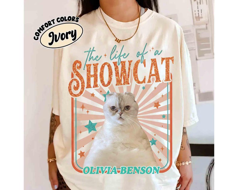 Może przedstawiać: Kremowa koszulka z napisem "the life of a SHOWCAT" w kolorze pomarańczowym i brokatowymi literami. Ilustracja kota jest wyśrodkowana, z retro wzorem z gwiazdami i paskami. Poniżej znajduje się imię "OLIVIA-BENSON".