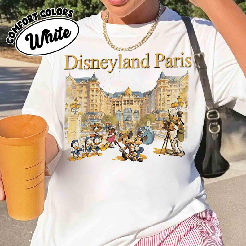 May include: White t-shirt with a Disneyland Paris graphic featuring cartoon characters and a detailed building facade. The shirt has the words "COMFORT COLORS" and "White" in a circular design at the top left.
