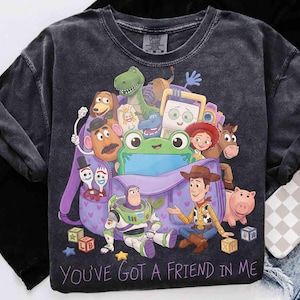 May include: A charcoal gray t-shirt featuring a graphic print of Toy Story characters including Woody, Buzz Lightyear, and a green frog emerging from a purple backpack. The text "YOU'VE GOT A FRIEND IN ME" is printed in purple at the bottom of the design.
