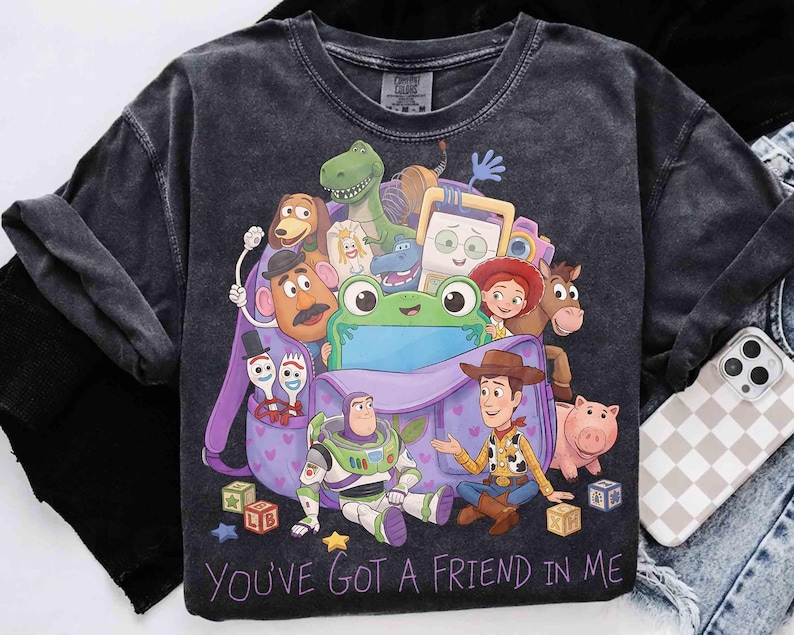 May include: A charcoal gray t-shirt featuring a graphic print of Toy Story characters including Woody, Buzz Lightyear, and a green frog emerging from a purple backpack. The text "YOU'VE GOT A FRIEND IN ME" is printed in purple at the bottom of the design.