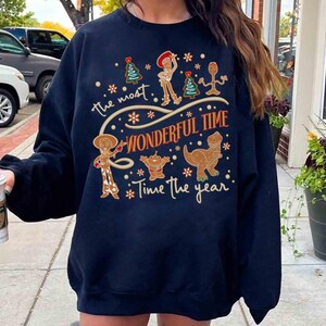 Puede incluir: Sudadera azul marino con un dise&ntilde;o festivo con personajes de dibujos animados y el texto "the most wonderful time of the year". El dise&ntilde;o incluye un &aacute;rbol de Navidad, un vaquero, un extraterrestre y un dinosaurio.