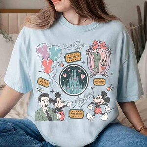 Peut inclure: T-shirt bleu clair avec un motif graphique color&eacute; repr&eacute;sentant Mickey Mouse, un ch&acirc;teau, des ballons et des billets de parc. Le motif comprend le texte "Walt Disney" et "Mickey Mouse" dans un style fantaisiste.