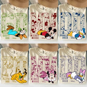 May include: Cream-colored t-shirts featuring Disney characters. Each shirt has a large character name and image: Goofy, Mickey Mouse, Donald Duck, Pluto, Minnie Mouse, and Daisy Duck. The shirts have a collegiate style design.