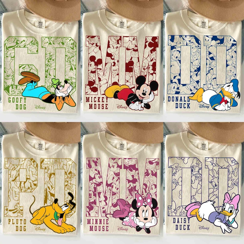 May include: Cream-colored t-shirts featuring Disney characters. Each shirt has a large character name and image: Goofy, Mickey Mouse, Donald Duck, Pluto, Minnie Mouse, and Daisy Duck. The shirts have a collegiate style design.
