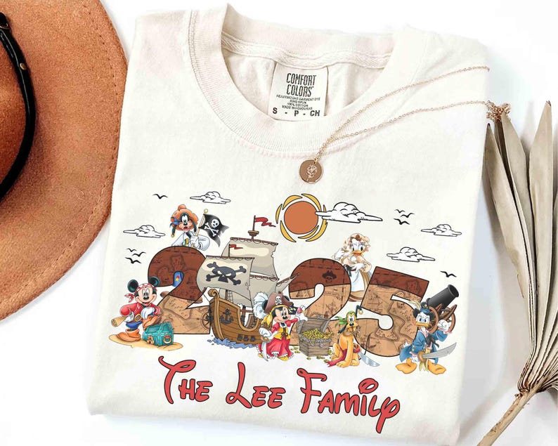 May include: Off-white t-shirt with a pirate-themed design featuring cartoon characters, the number "25", and the text "The Lee Family" in red. A gold necklace and a brown hat are also visible.