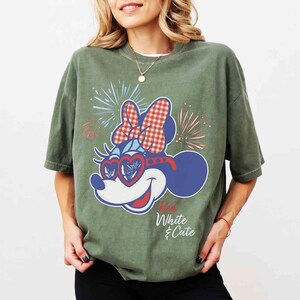Op de afbeelding: Groen t-shirt met een Minnie Mouse-afbeelding met een rood-witte ruitjesstrik, hartvormige zonnebril en vuurwerk. De tekst "Red, White & Cute" is eronder gedrukt. Het shirt heeft korte mouwen en een ronde hals.