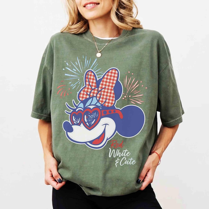 Op de afbeelding: Groen t-shirt met een Minnie Mouse-afbeelding met een rood-witte ruitjesstrik, hartvormige zonnebril en vuurwerk. De tekst "Red, White & Cute" is eronder gedrukt. Het shirt heeft korte mouwen en een ronde hals.
