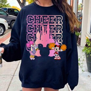 May include: Black sweatshirt featuring the word "CHEER" in pink block letters, with a pink castle silhouette. Cartoon characters Daisy Duck and Minnie Mouse are also featured, with Daisy holding pom-poms and Minnie in a cheerleader outfit.