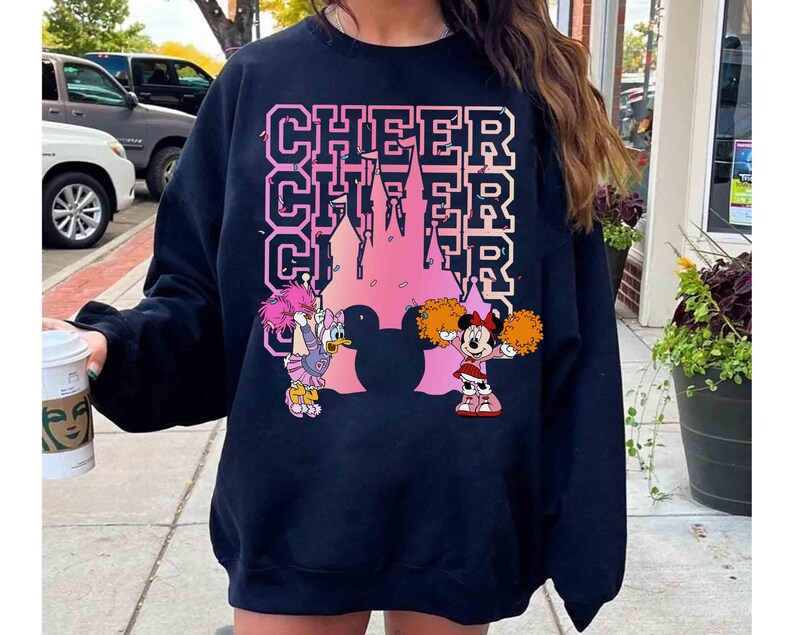 May include: Black sweatshirt featuring the word "CHEER" in pink block letters, with a pink castle silhouette. Cartoon characters Daisy Duck and Minnie Mouse are also featured, with Daisy holding pom-poms and Minnie in a cheerleader outfit.
