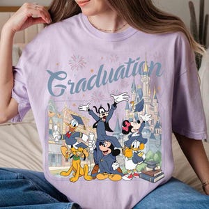 May include: A light purple t-shirt featuring a graphic of Disney characters in graduation attire. The word "Graduation" is written in a script font above the characters. The characters include Mickey Mouse, Minnie Mouse, Donald Duck, Goofy, and Pluto.