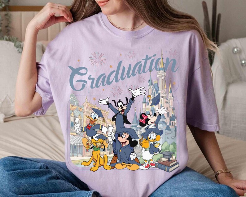 May include: A light purple t-shirt featuring a graphic of Disney characters in graduation attire. The word "Graduation" is written in a script font above the characters. The characters include Mickey Mouse, Minnie Mouse, Donald Duck, Goofy, and Pluto.