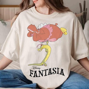 May include: Cream-colored t-shirt featuring a cartoon illustration from Disney's Fantasia. The design includes a pink hippo, a yellow and green alligator, and the word "Fantasia" in black. The shirt is a relaxed fit.