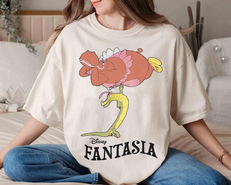 May include: Cream-colored t-shirt featuring a cartoon illustration from Disney's Fantasia. The design includes a pink hippo, a yellow and green alligator, and the word "Fantasia" in black. The shirt is a relaxed fit.