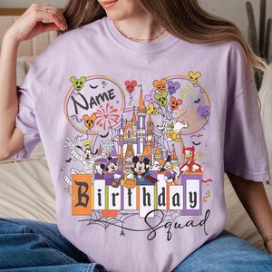 May include: A lilac-colored t-shirt featuring a colorful graphic of a castle, balloons, and cartoon characters. The text "Name" is above the castle, and "Birthday Squad" is below. The design includes fireworks and spiderwebs.