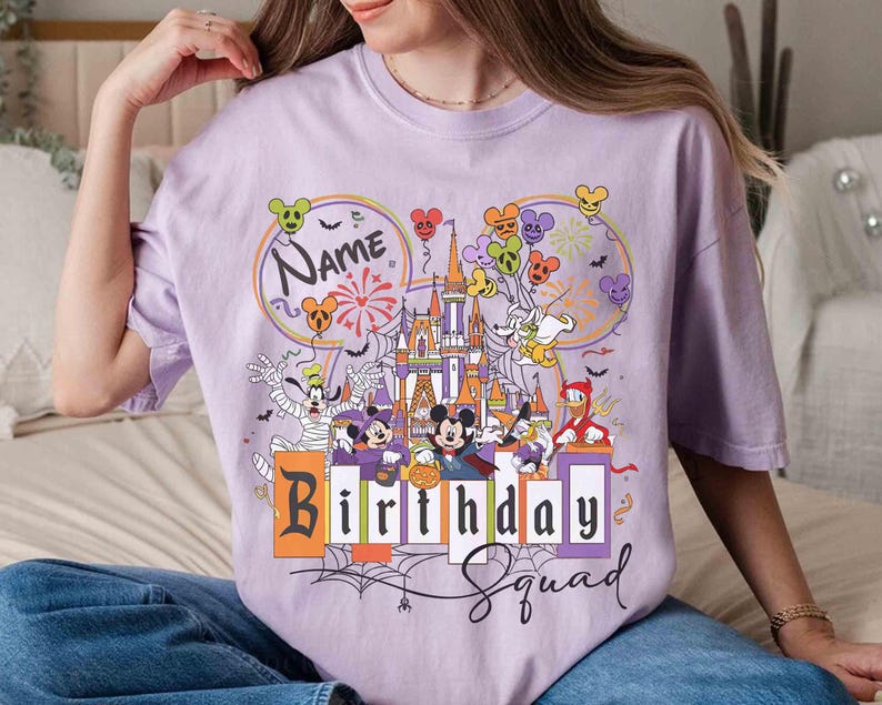 May include: A lilac-colored t-shirt featuring a colorful graphic of a castle, balloons, and cartoon characters. The text "Name" is above the castle, and "Birthday Squad" is below. The design includes fireworks and spiderwebs.