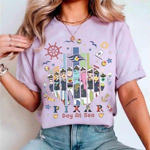 K&ouml;nnte beinhalten: Ein lilafarbenes T-Shirt mit einer Grafik von Pixar-Charakteren in Matrosenuniformen. Das Design umfasst ein Steuerrad, Sterne und den Text "PIXAR Day At Sea" in bunten Buchstaben. Das Shirt hat hochgekrempelte &Auml;rmel.