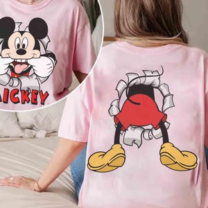 May include: Pink t-shirt featuring a graphic of Mickey Mouse bursting through a torn paper effect. The front of the shirt shows Mickey's face with the word "Mickey" below. The back of the shirt shows Mickey's legs and yellow shoes.
