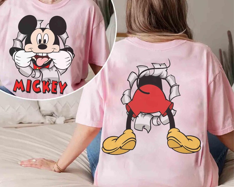 May include: Pink t-shirt featuring a graphic of Mickey Mouse bursting through a torn paper effect. The front of the shirt shows Mickey's face with the word "Mickey" below. The back of the shirt shows Mickey's legs and yellow shoes.